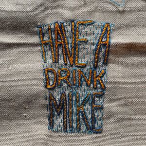 "Have A Drink Mike" embroidered in multicoloured thread on tabby weave linen in the shape of a glass