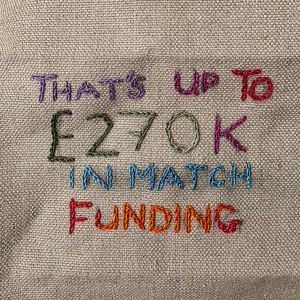 "That's up to "270K in match funding" embroidered in multicoloured thread on tabby weave linen